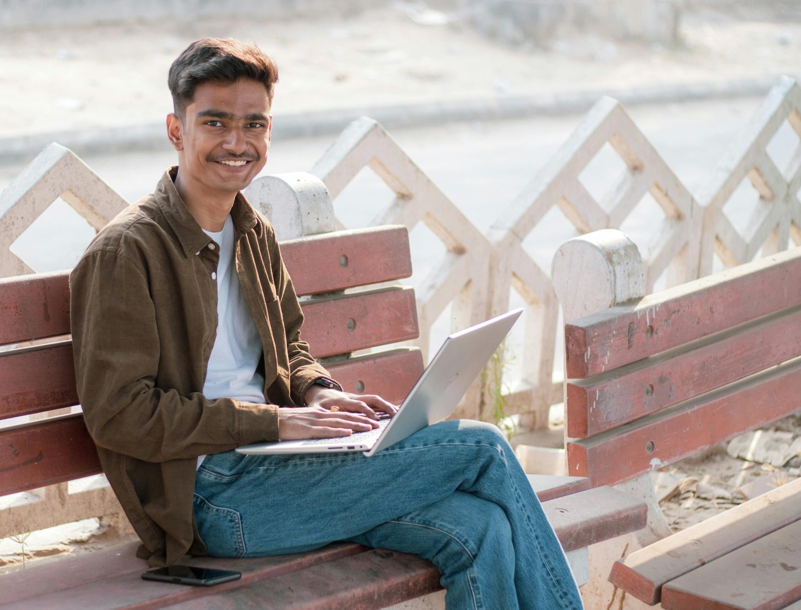 Smiling young adult using a laptop while sitting on a park bench outdoors.