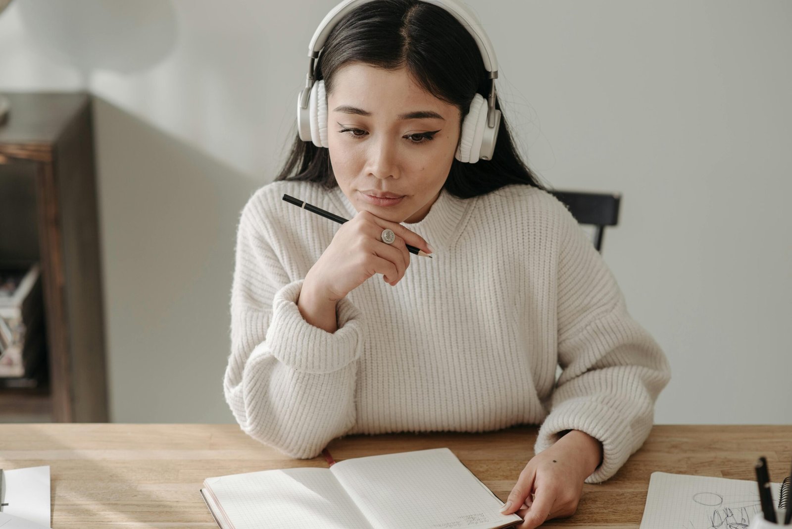 An Asian woman in a white sweater writing in a notebook with headphones on, sitting at a wooden table indoors.